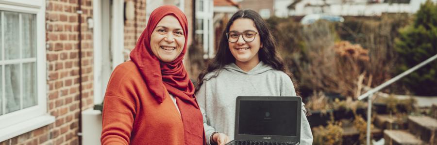 A mother and daughter stand in their front garden facing the camera and smiling. The young daughter is holding a silver laptop.