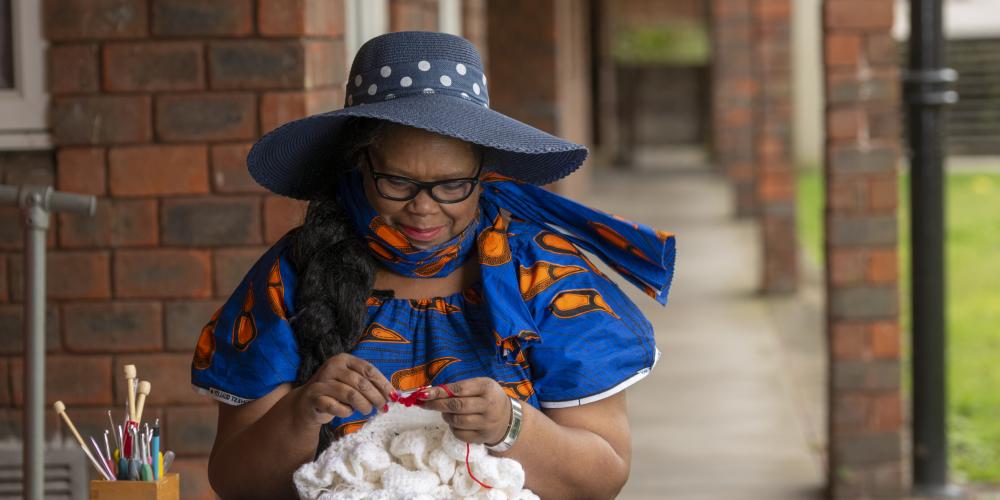 A lady wearing a hat, sits at a table doing crochet